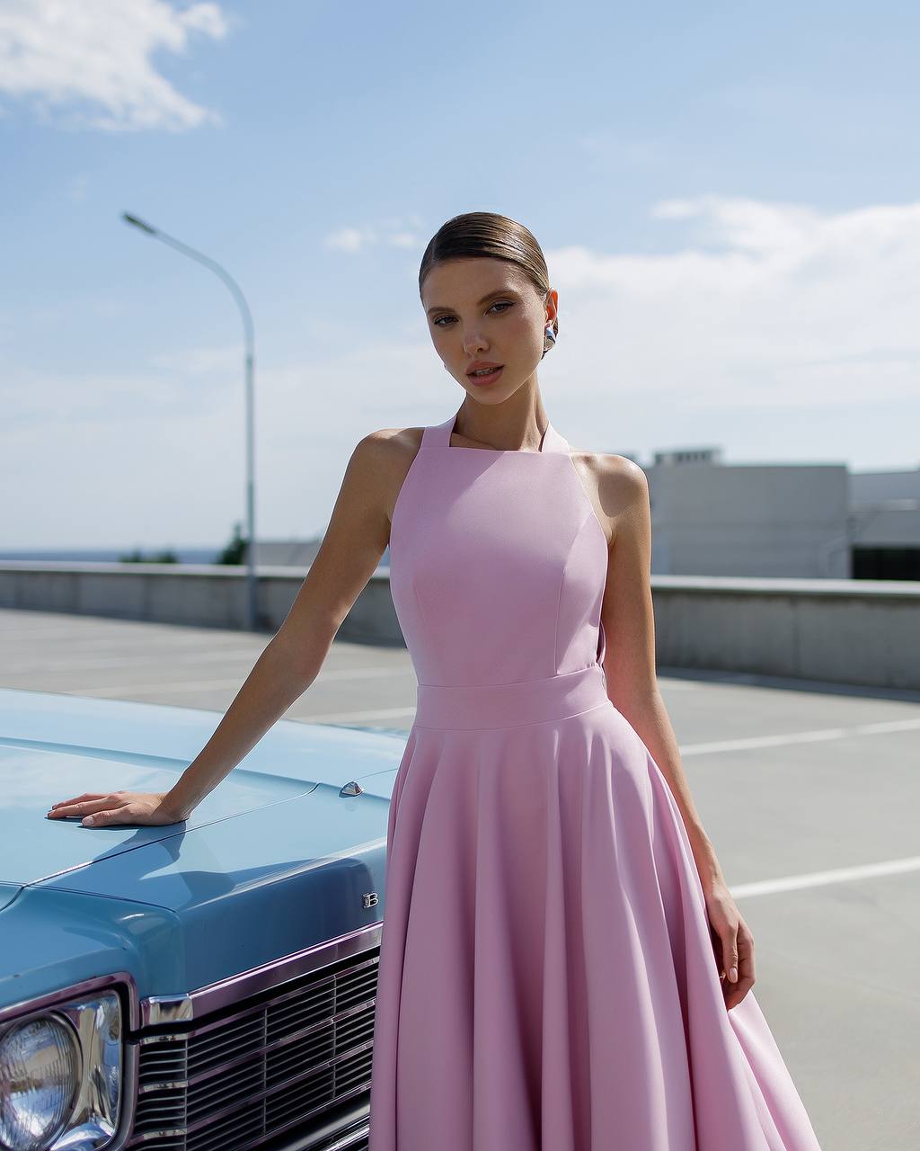 Woman in a pink dress standing next to a blue car on a clear day.