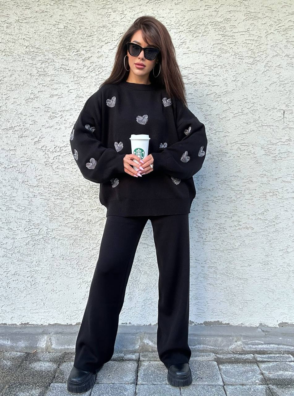 Person wearing a black sweater with heart patterns and holding a Starbucks cup against a light gray wall.