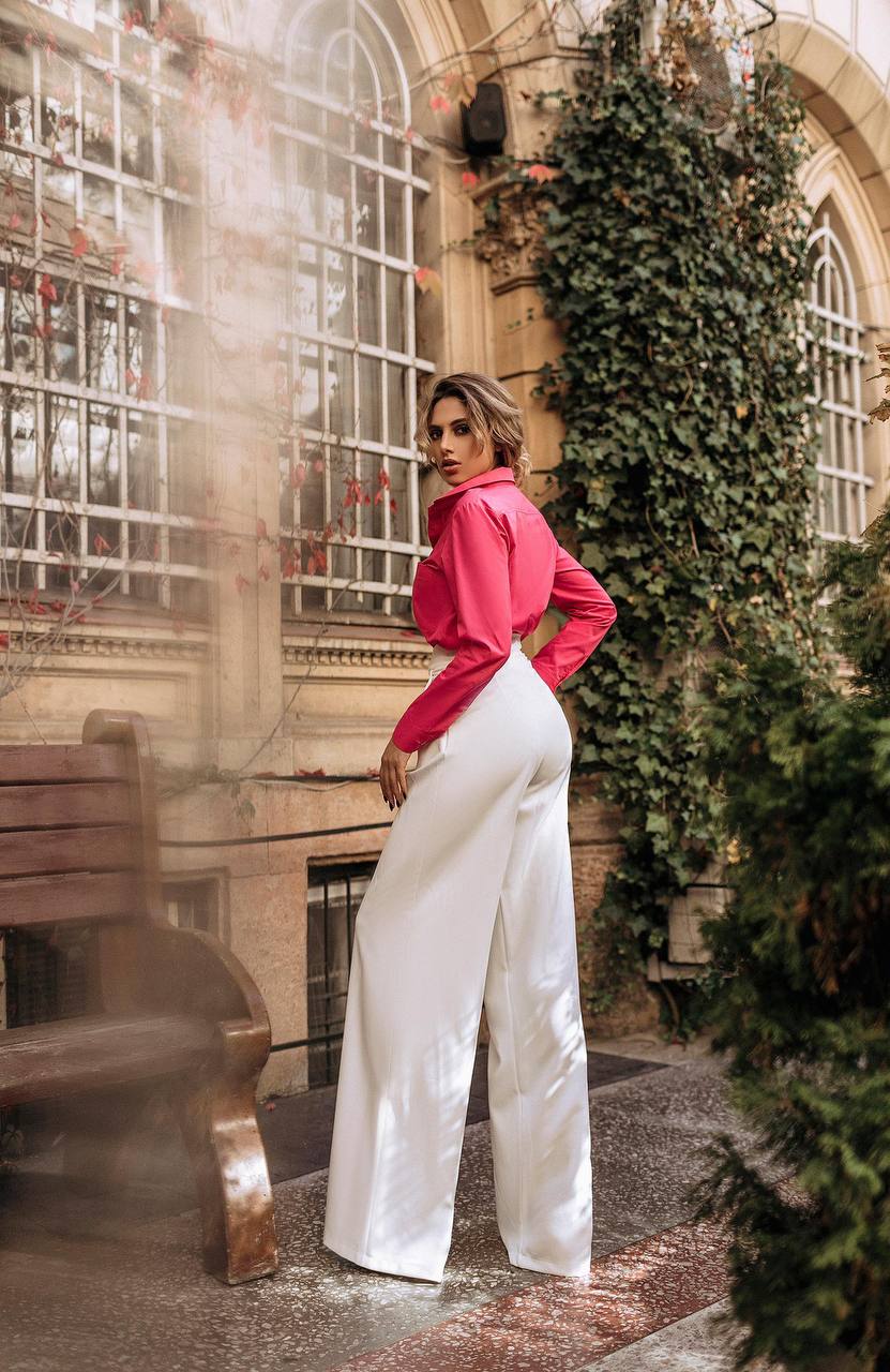 Woman in a pink blouse and white pants standing in an outdoor setting with greenery and a bench.