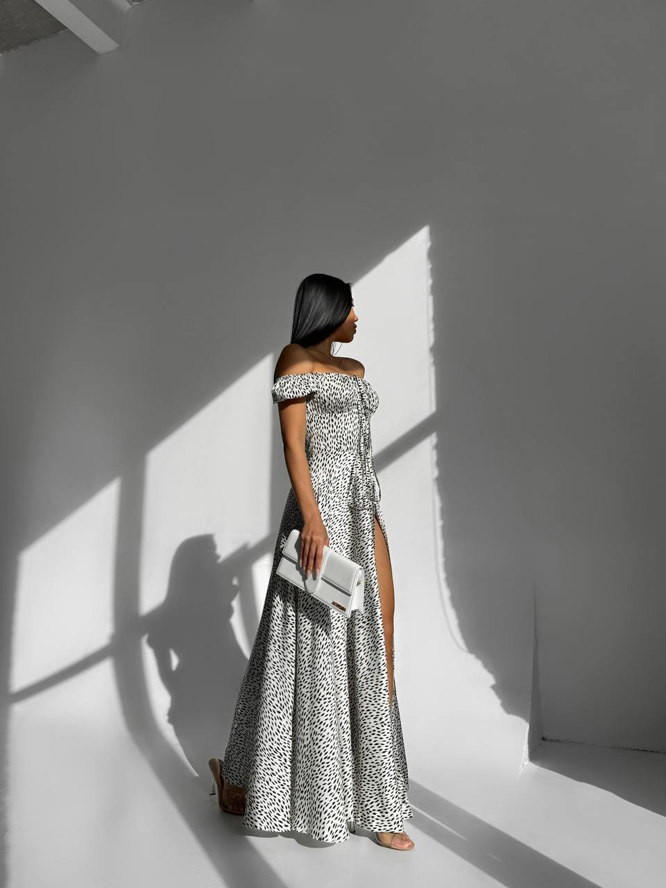 Woman in a long, off-shoulder dress with a pattern standing against a white wall with light and shadow play.