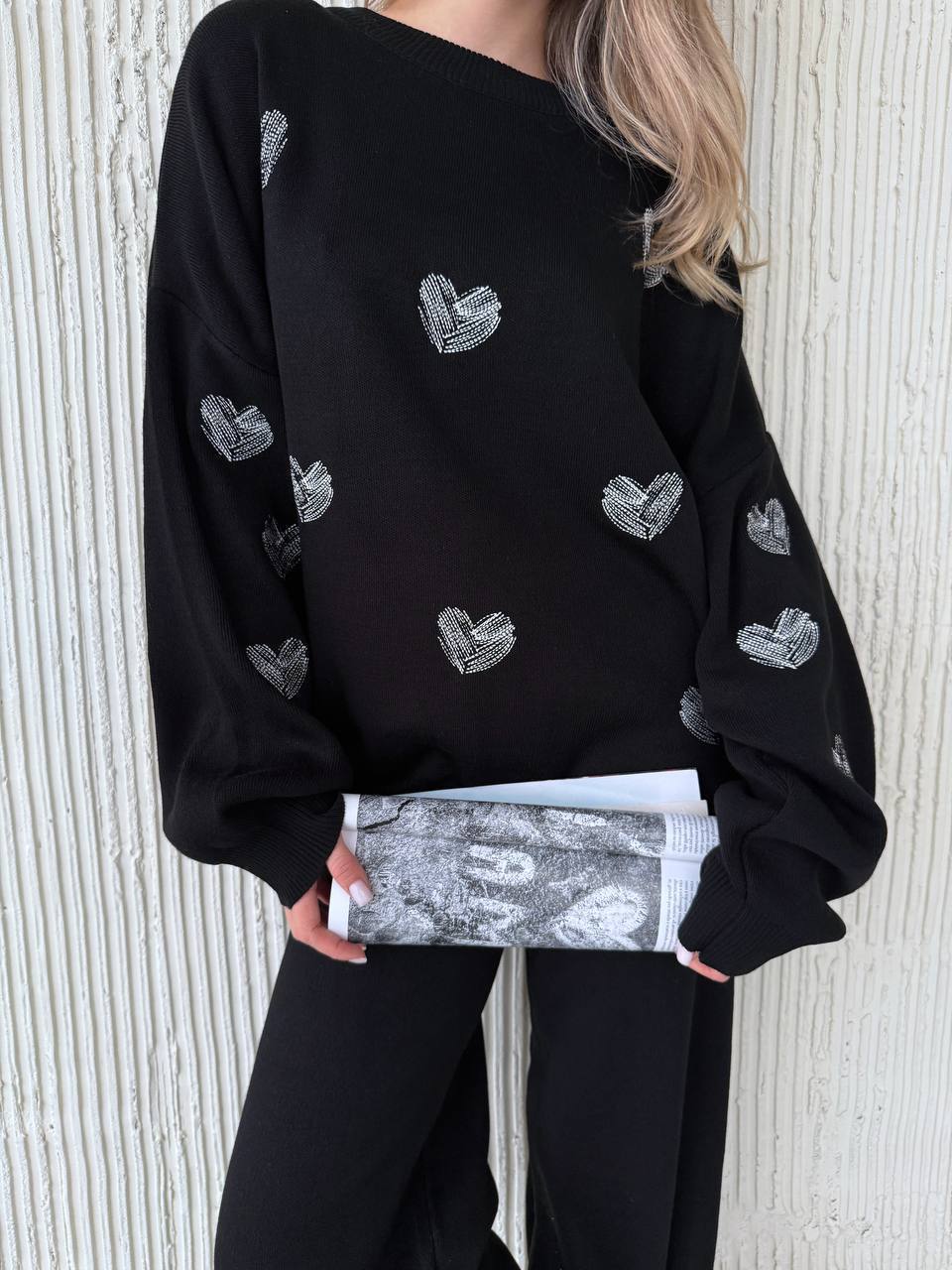 Person wearing a black sweater with heart patterns, holding a matching clutch against a light background