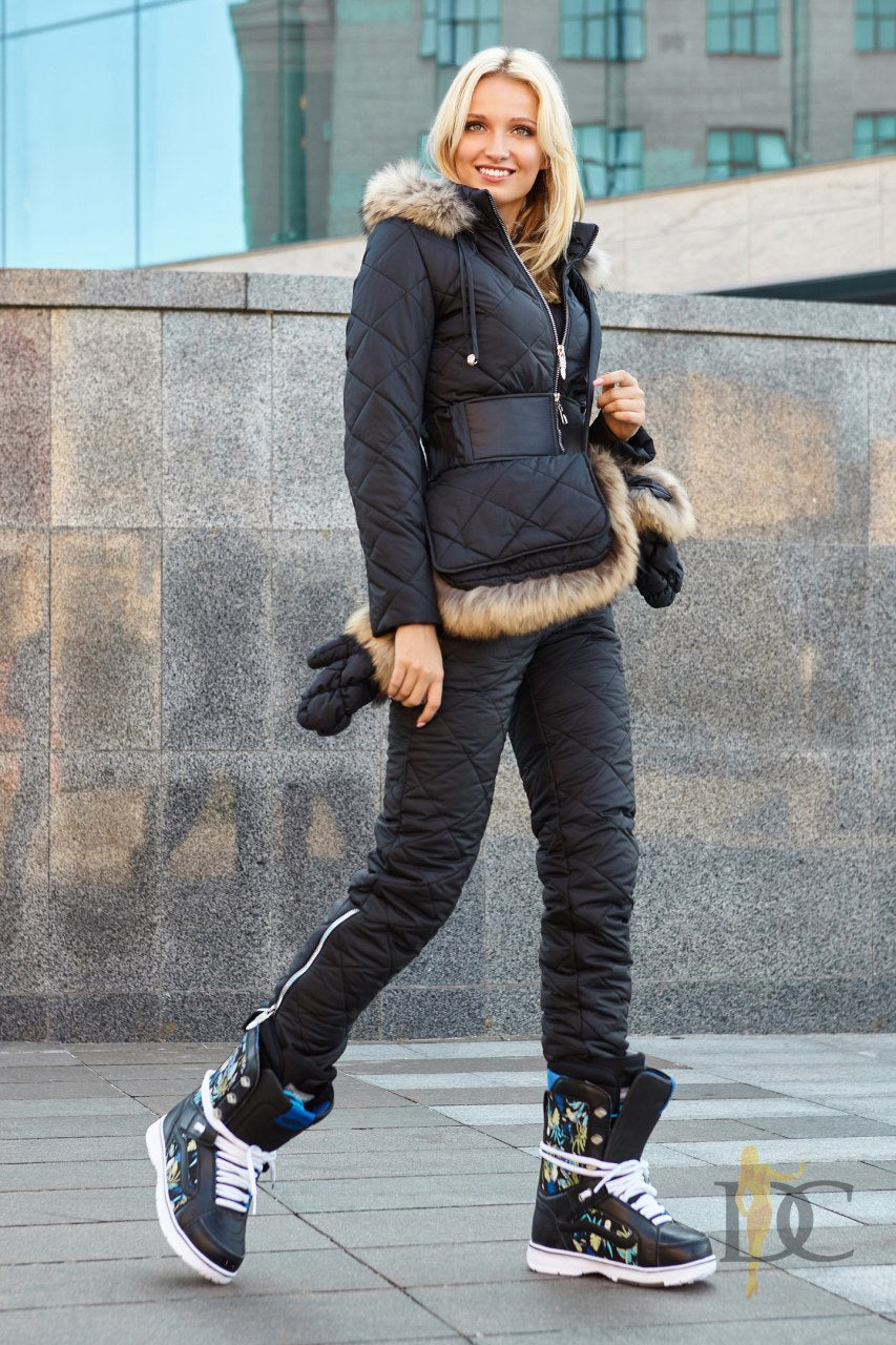 Woman wearing a black quilted jacket with fur trim and black pants, standing against a stone wall.