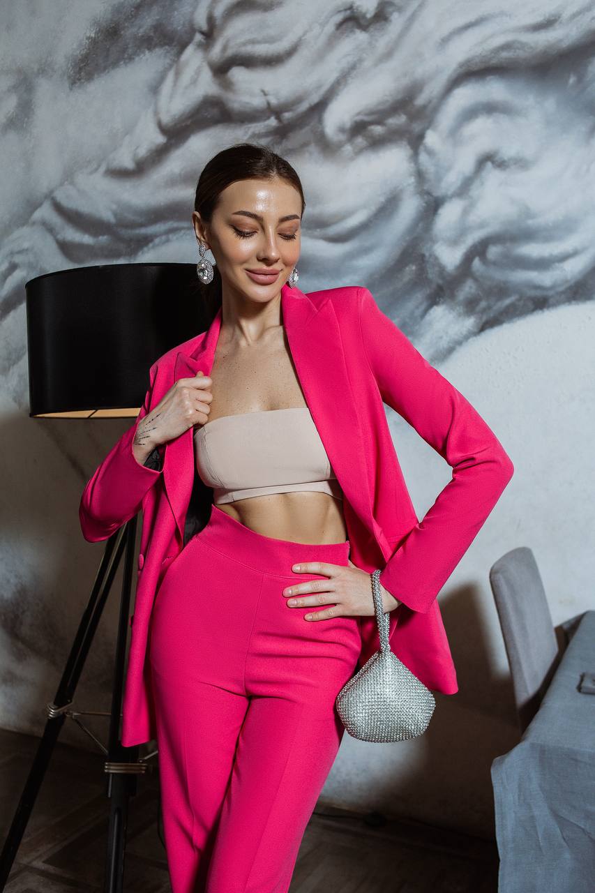 Woman in a bright pink suit posing indoors with a decorative wall in the background.