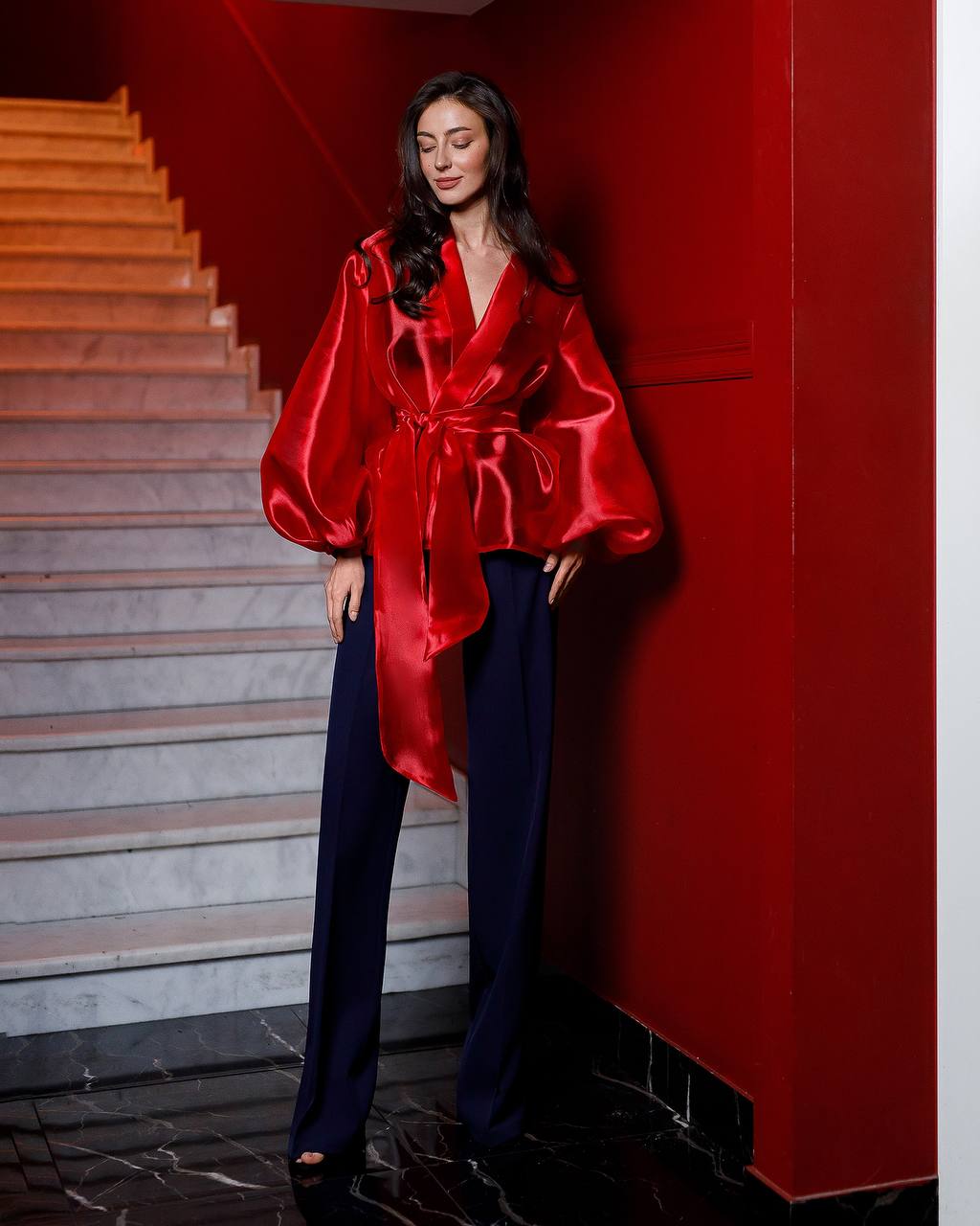 Woman in a red satin top and blue pants standing against a red wall.