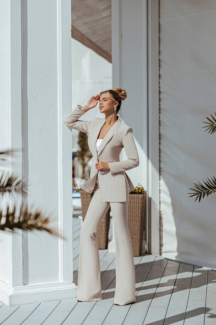 Woman in a beige suit standing in a sunlit room with white walls and wooden floor.