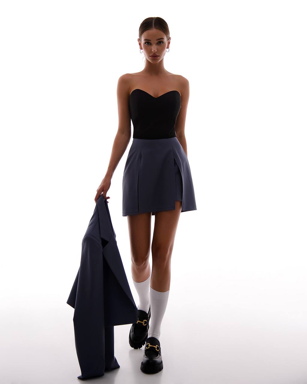 Woman in a black strapless top and gray skirt holding a navy blazer on a white background