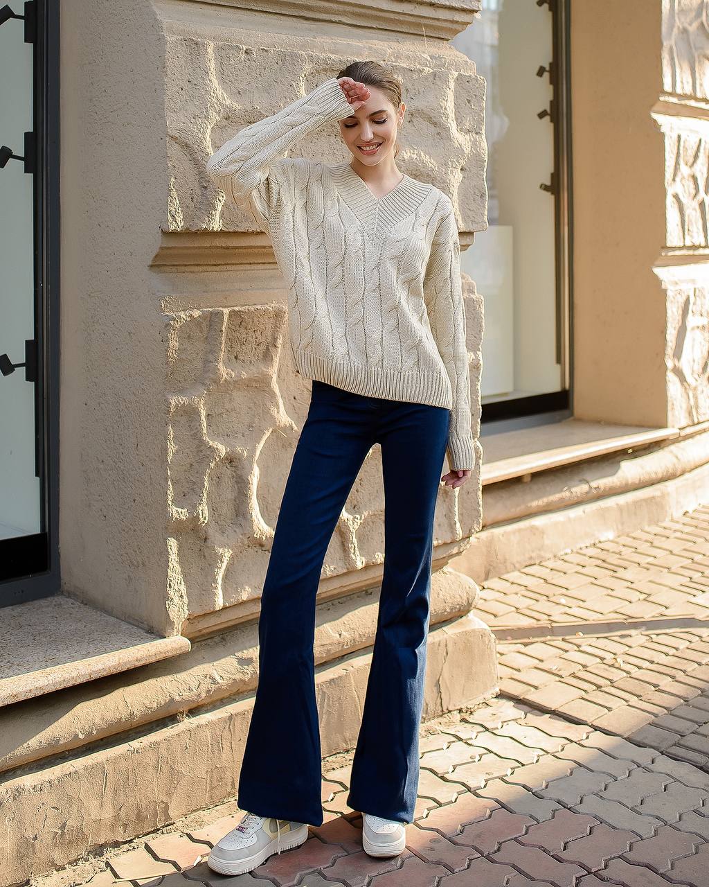 Woman wearing a cream sweater and blue jeans standing in front of a stone building.