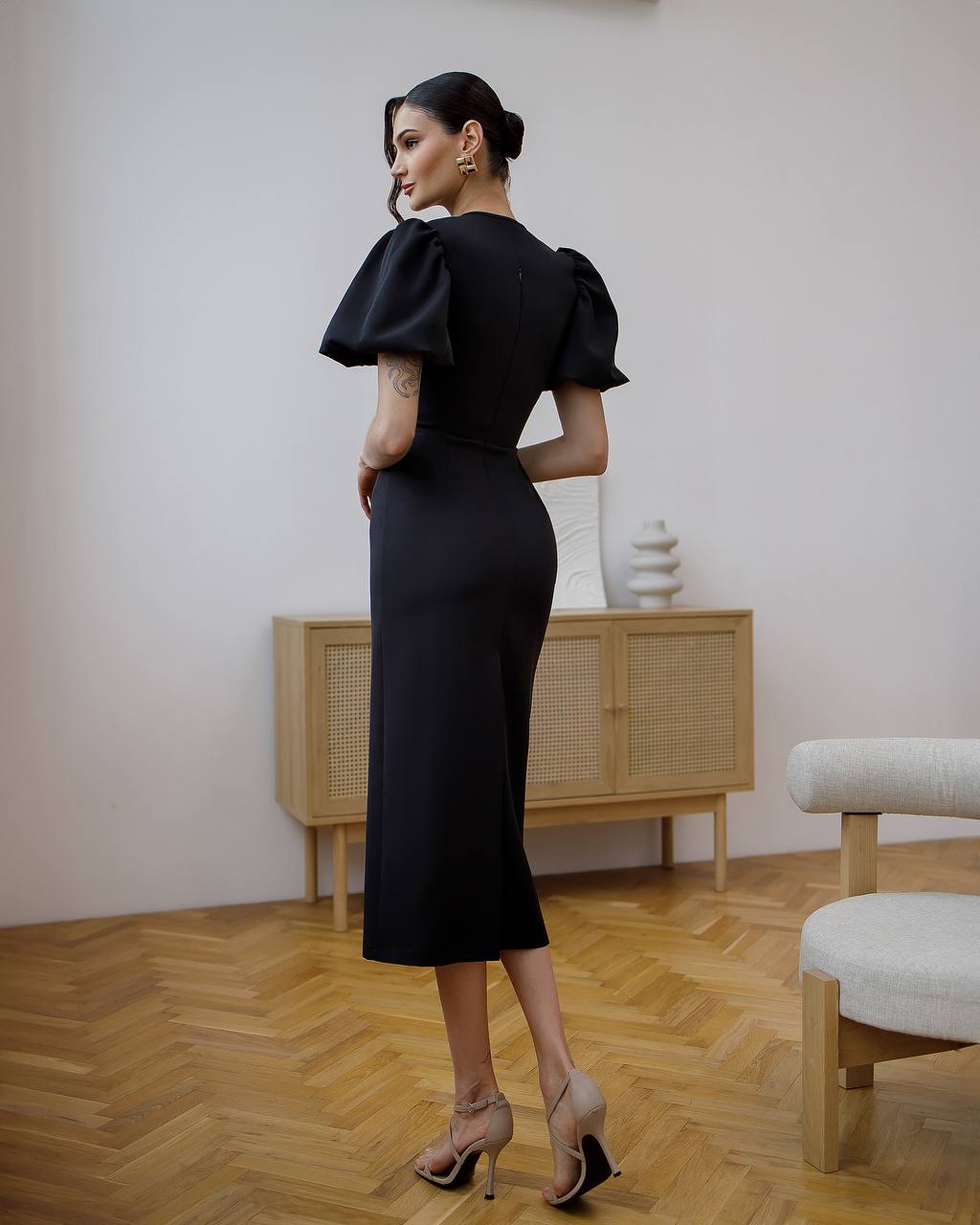 Woman in a black dress standing in a room with wooden flooring and a light-colored wall.