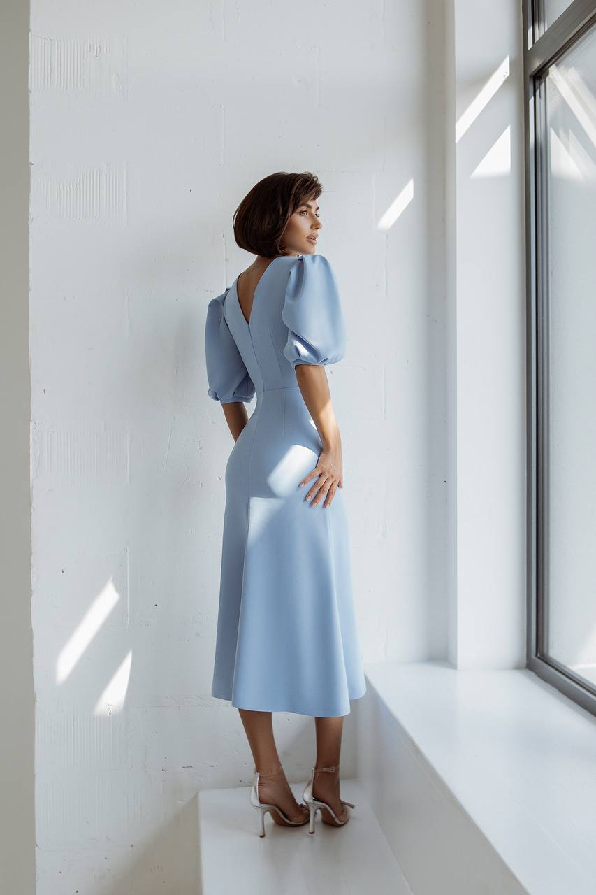 Woman in a light blue dress standing in a bright room with large windows.