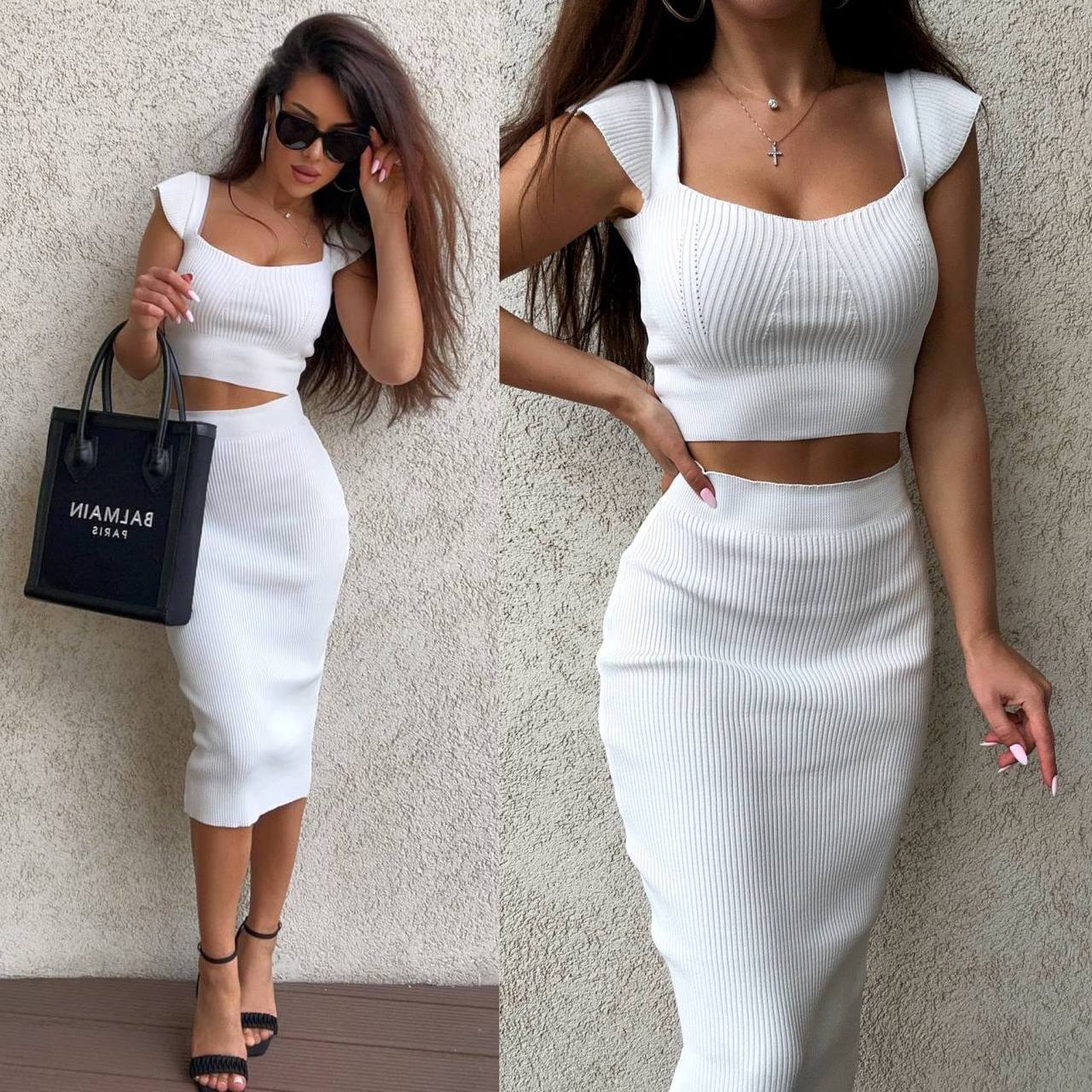 Woman wearing a white ribbed top and skirt with a black handbag against a neutral wall.