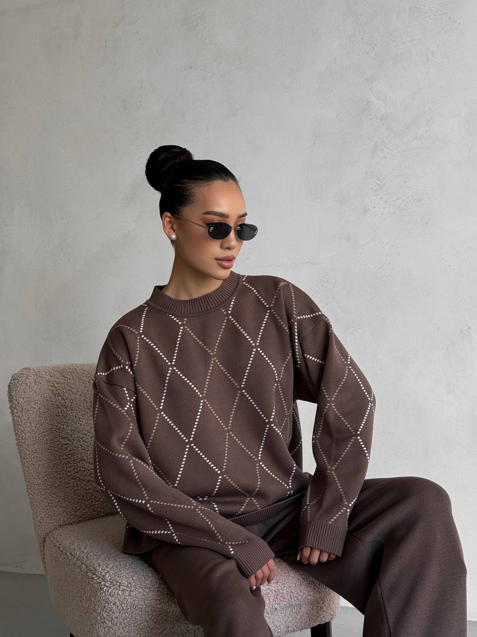 Person wearing a brown patterned sweater sitting on a chair against a plain background