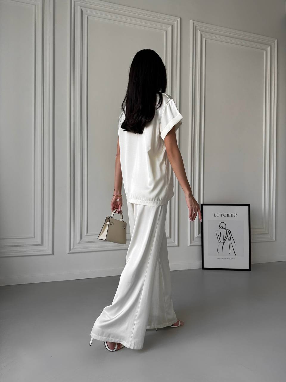 Woman in a white outfit standing in a room with a framed picture on the wall.