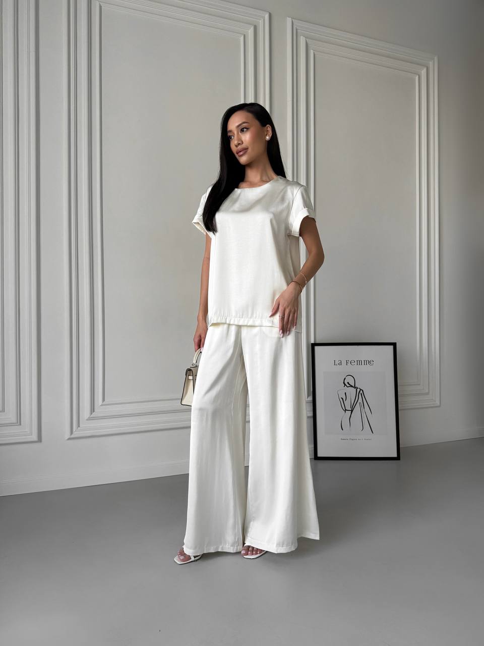 Woman in a white outfit standing against a gray wall with a framed picture.