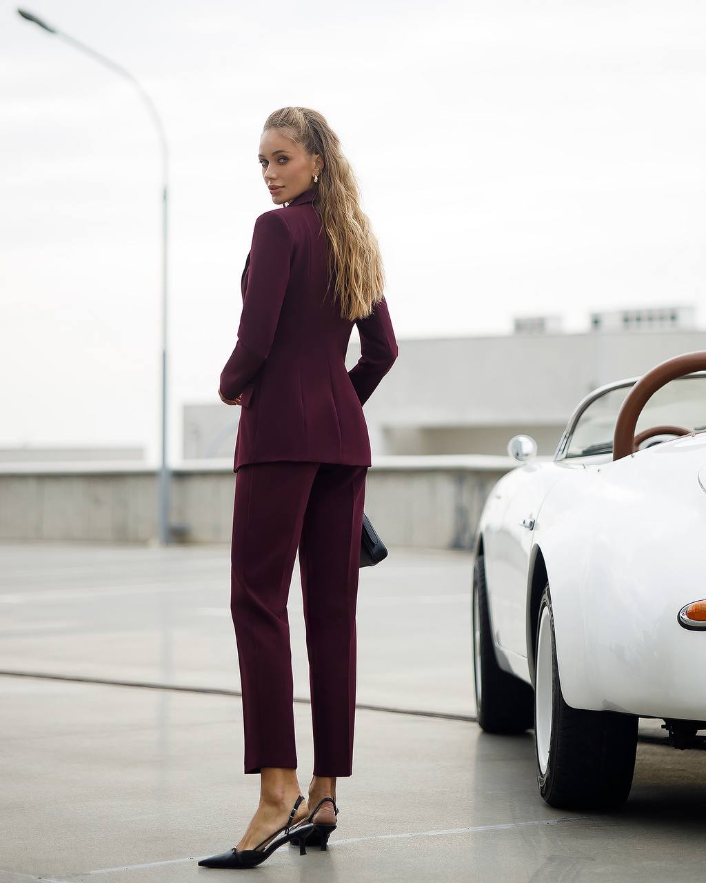 Woman in a burgundy suit standing next to a vintage car on a rooftop.
