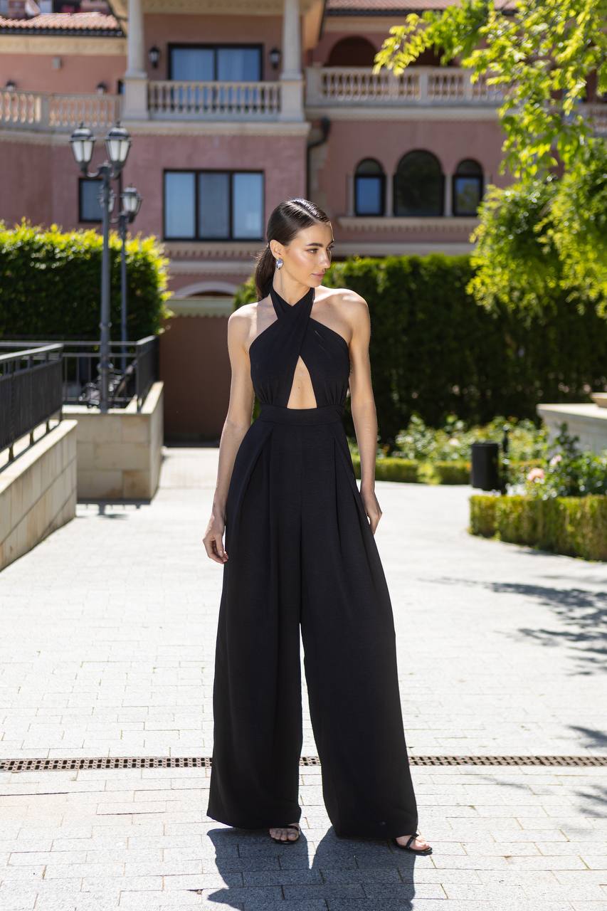 Woman in a black jumpsuit standing on a paved walkway with greenery and a building in the background