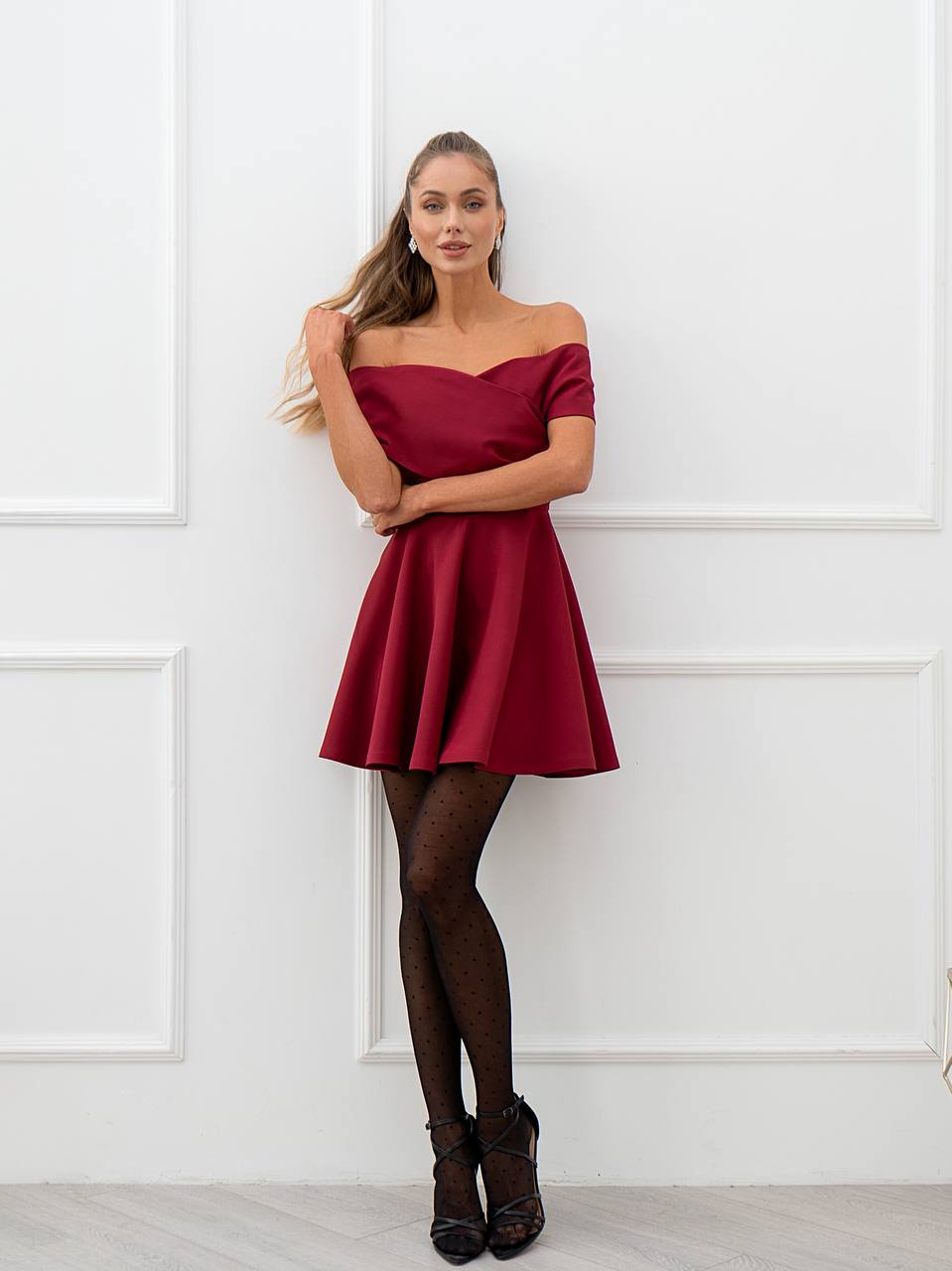 Woman wearing a red off-shoulder dress against a white paneled wall.