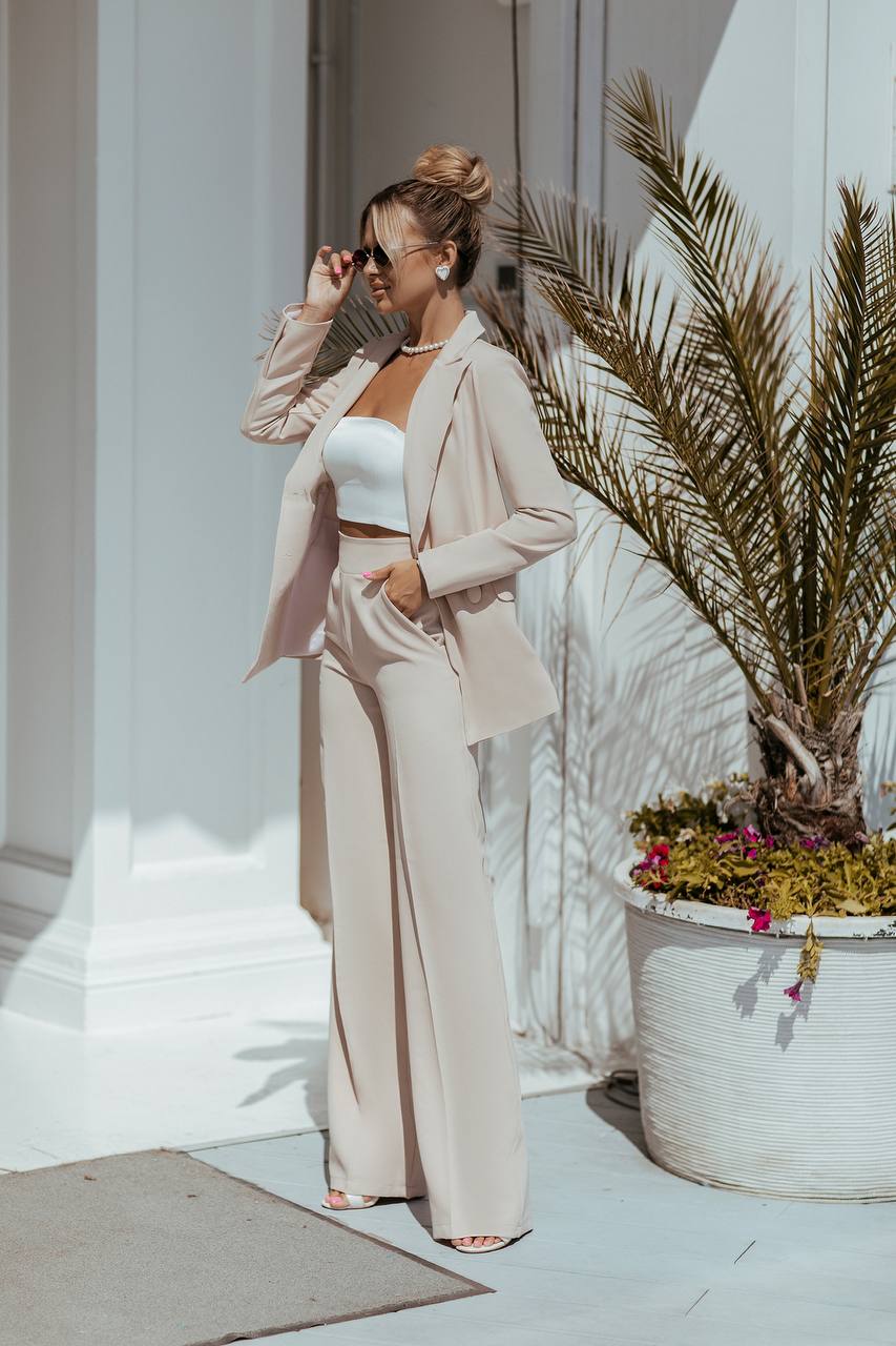 Woman in a beige suit standing next to a potted plant indoors