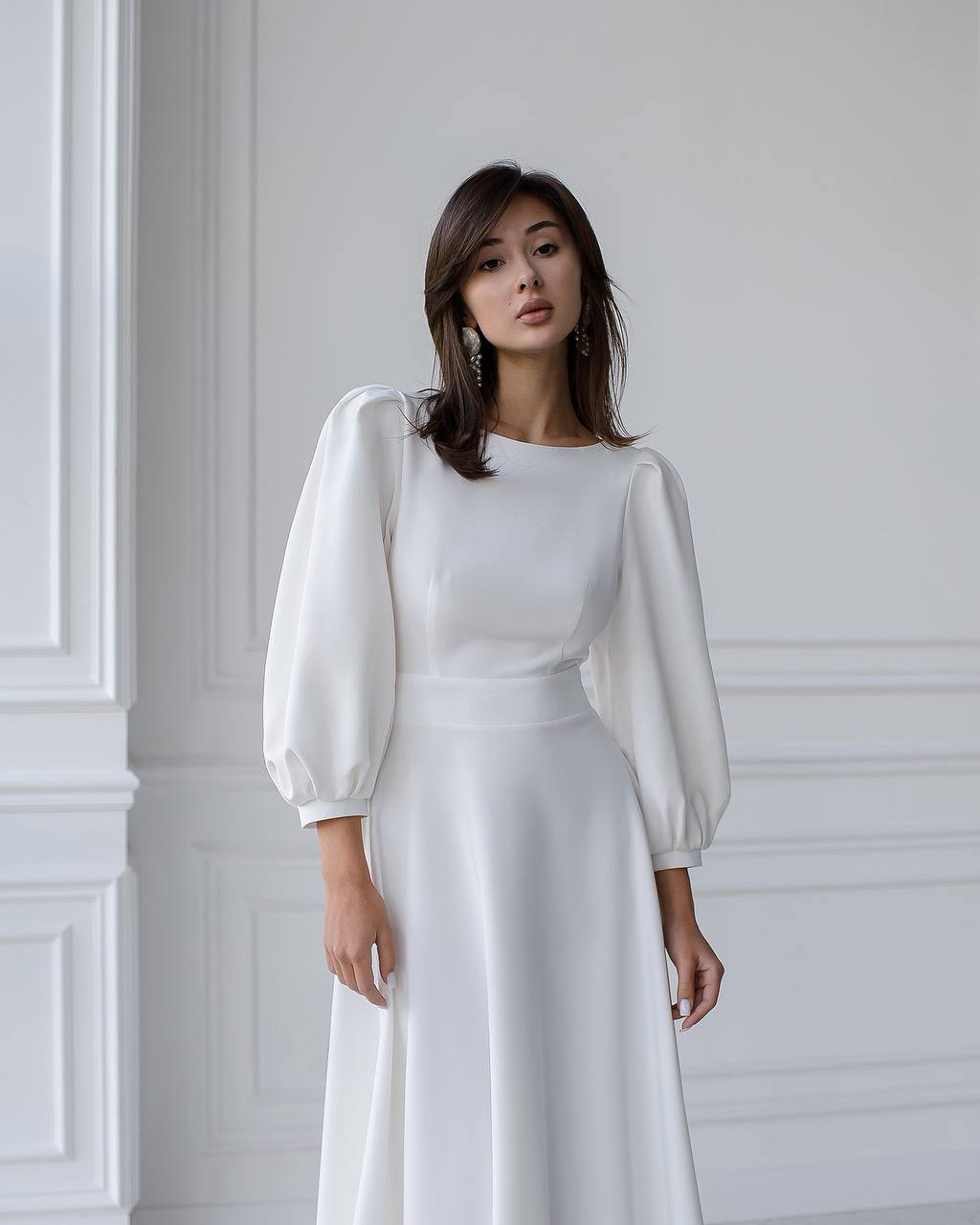 Woman wearing a white dress with sheer sleeves against a white wall