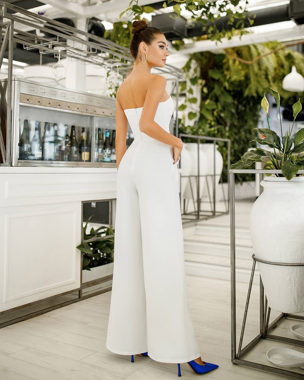 Woman in a white strapless jumpsuit standing in a modern indoor setting with plants.