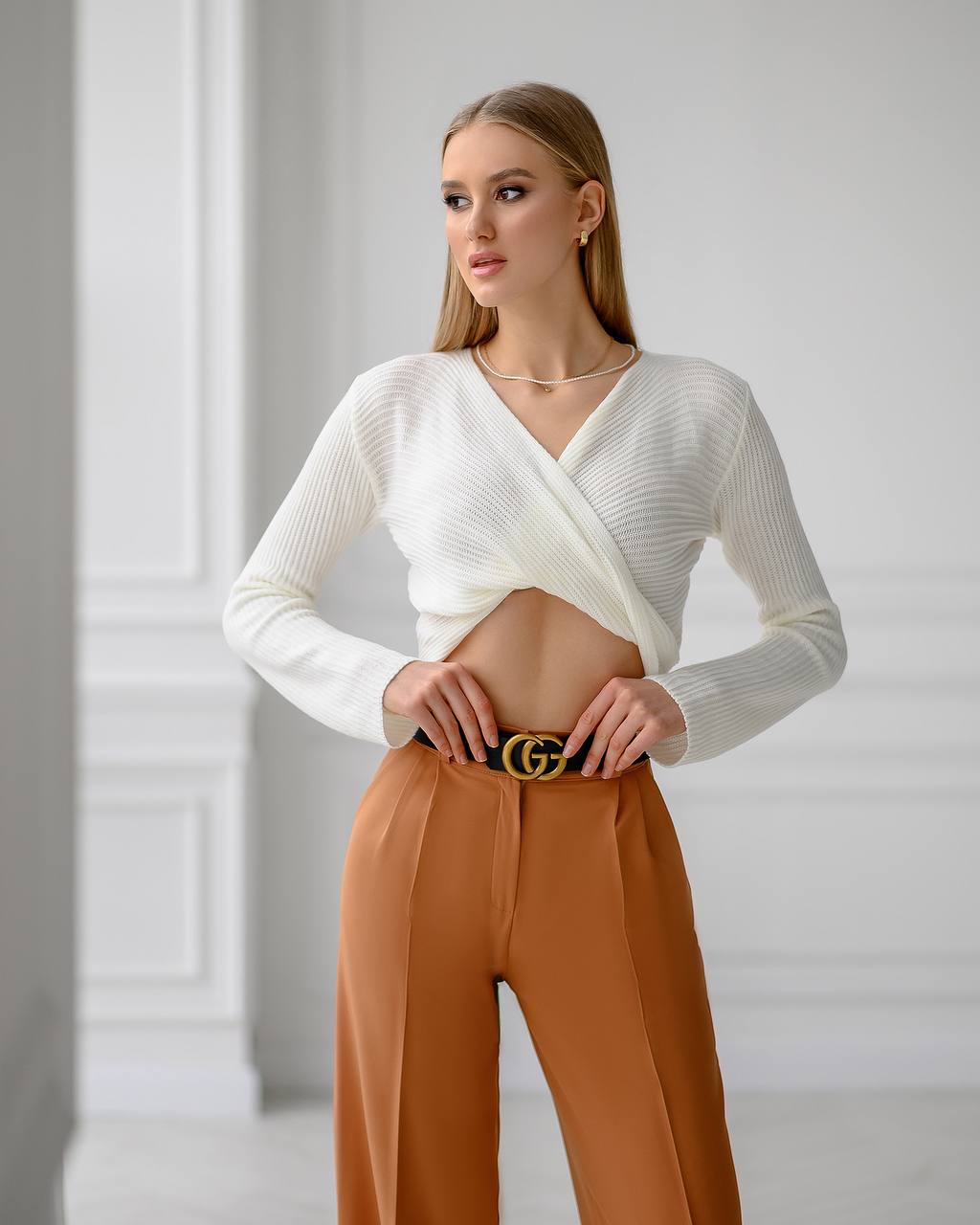 Woman wearing a white sweater and brown pants with a Gucci belt against a light gray background