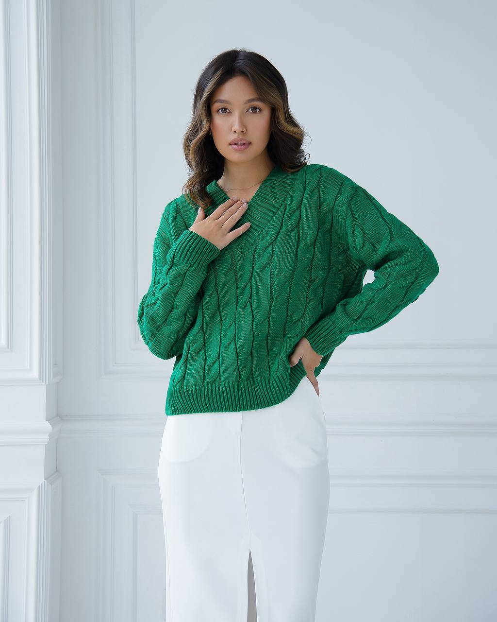 Woman wearing a green cable knit sweater and white pants against a light gray wall.