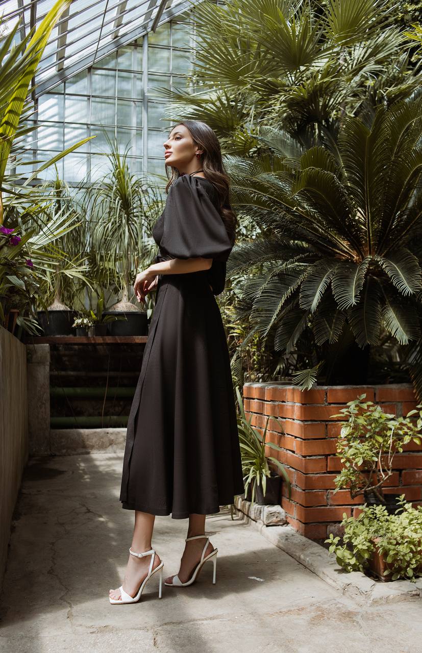 Woman in a black dress standing in a greenhouse filled with plants.
