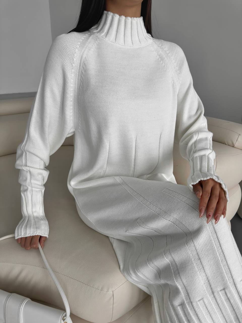 Person wearing a white ribbed knit sweater and pants sitting on a beige couch.