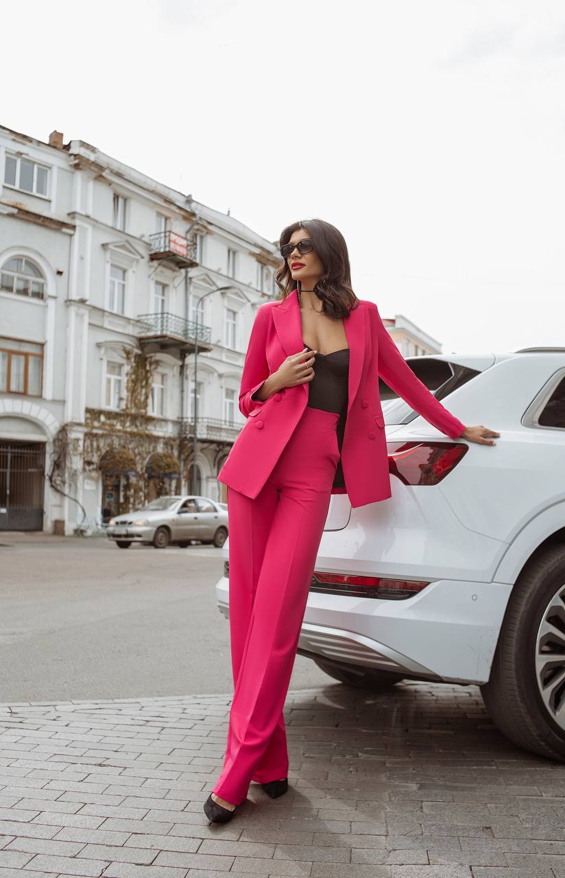 Woman in a bright pink suit standing next to a white car on a city street.