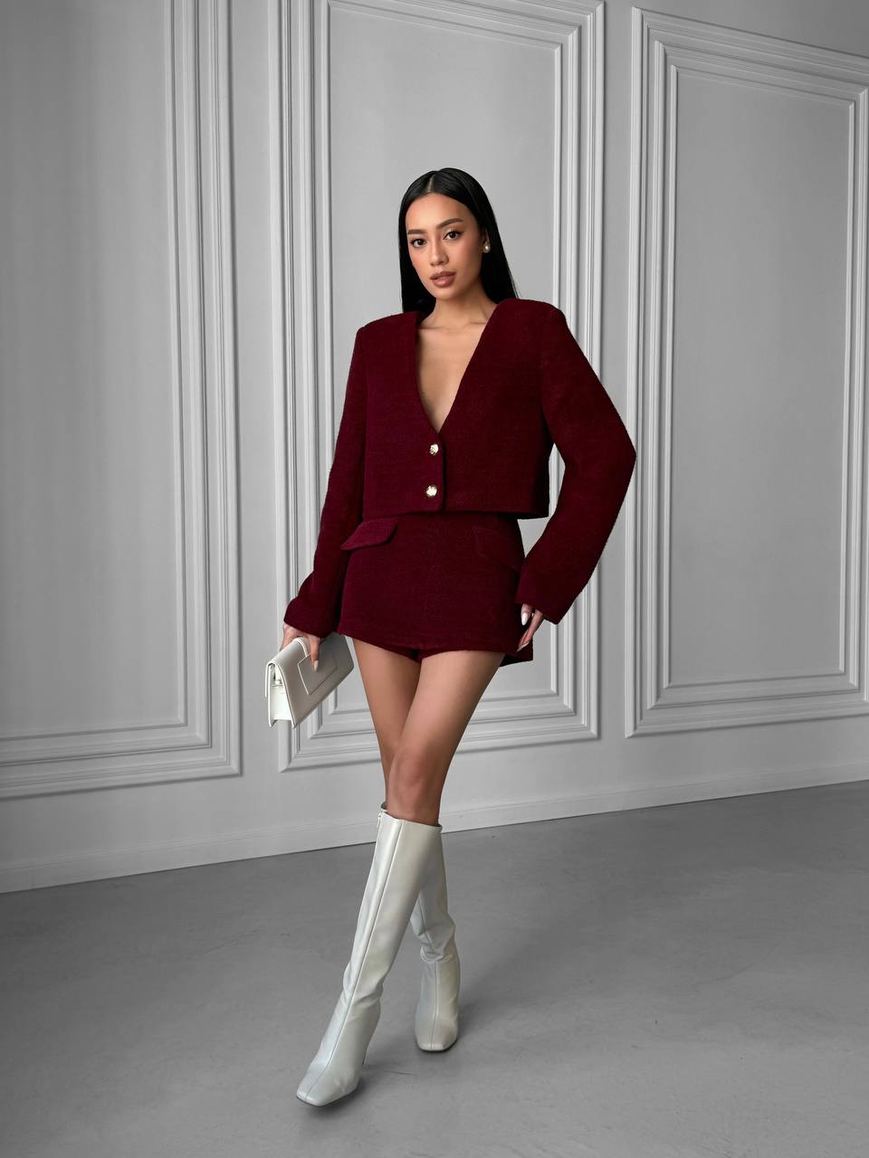 Woman in a burgundy outfit with white boots standing against a gray paneled wall.