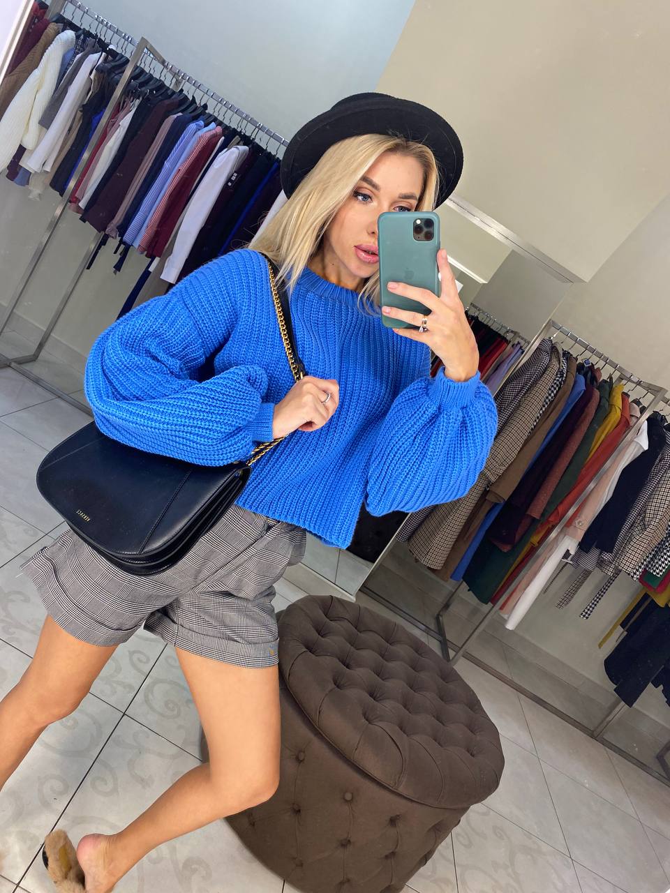 Person wearing a blue sweater and gray shorts taking a mirror selfie in a clothing store.