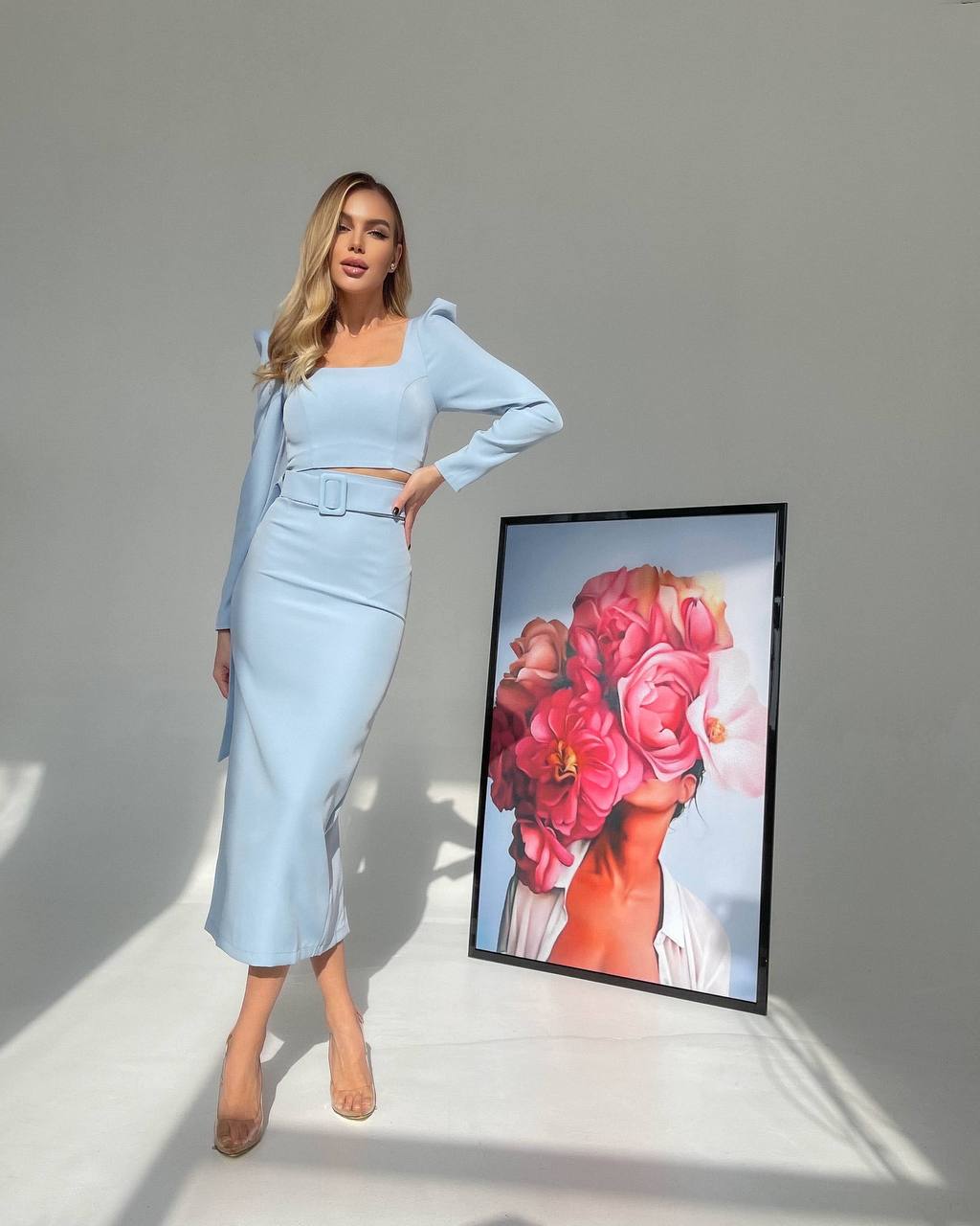 Woman in light blue outfit standing next to a framed artwork of a woman with flowers.