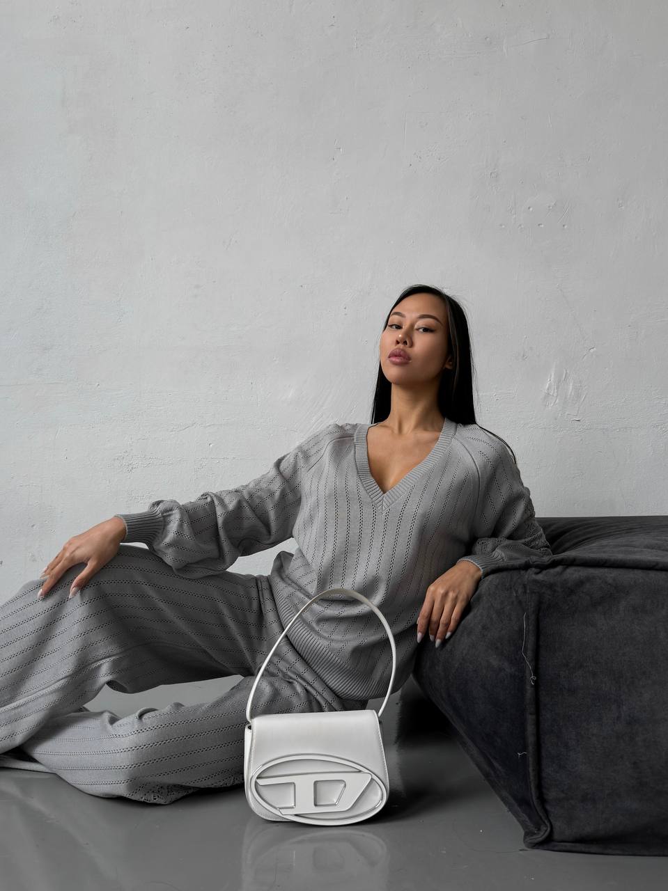 Woman in gray outfit sitting on a couch with a white handbag in front of her.