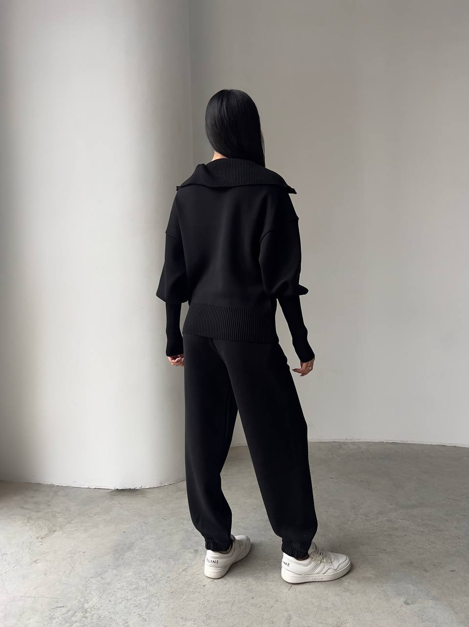 Person wearing a black outfit standing against a plain white wall.
