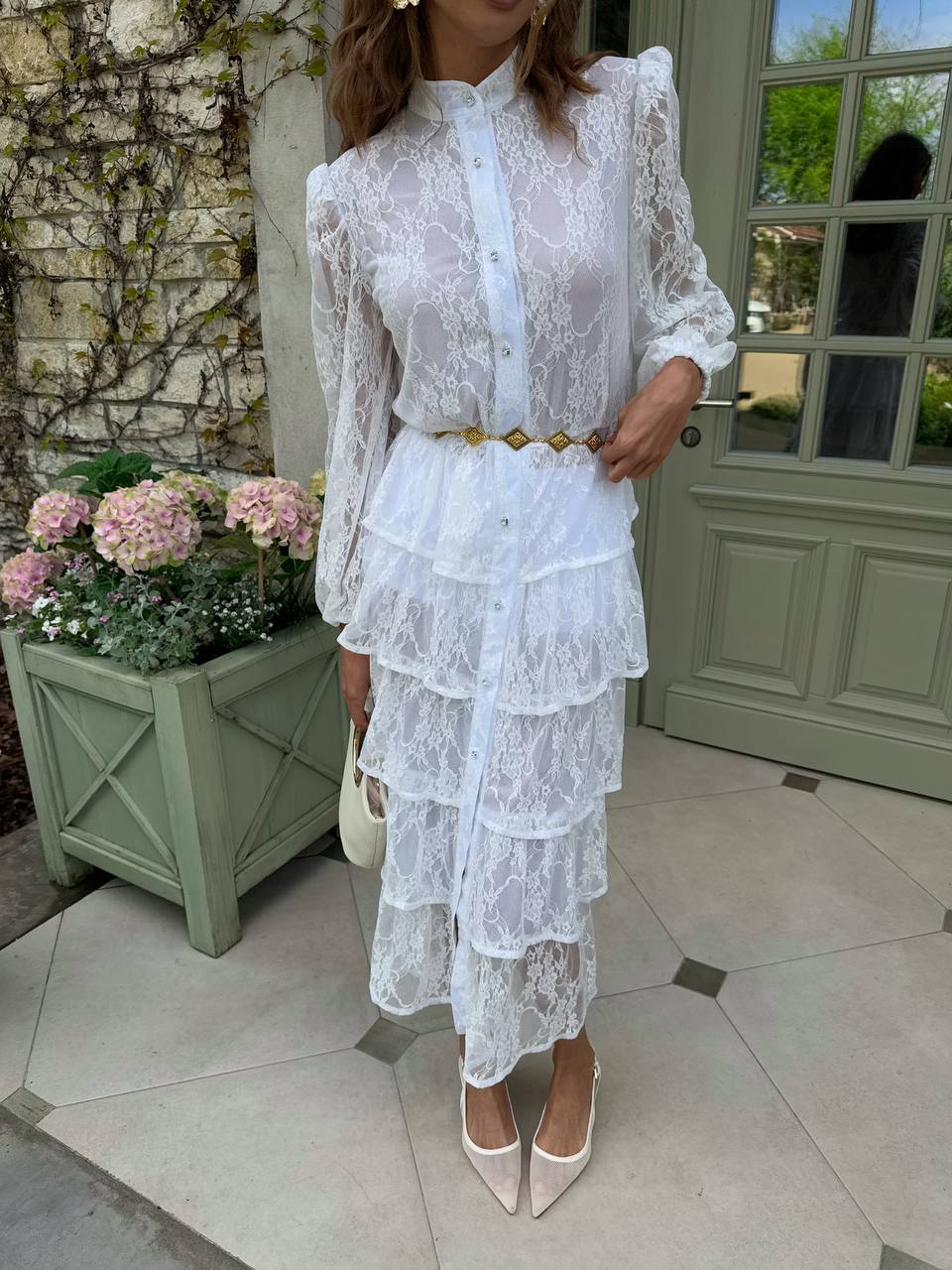 Woman wearing a white lace dress standing outdoors near a door and flowers.