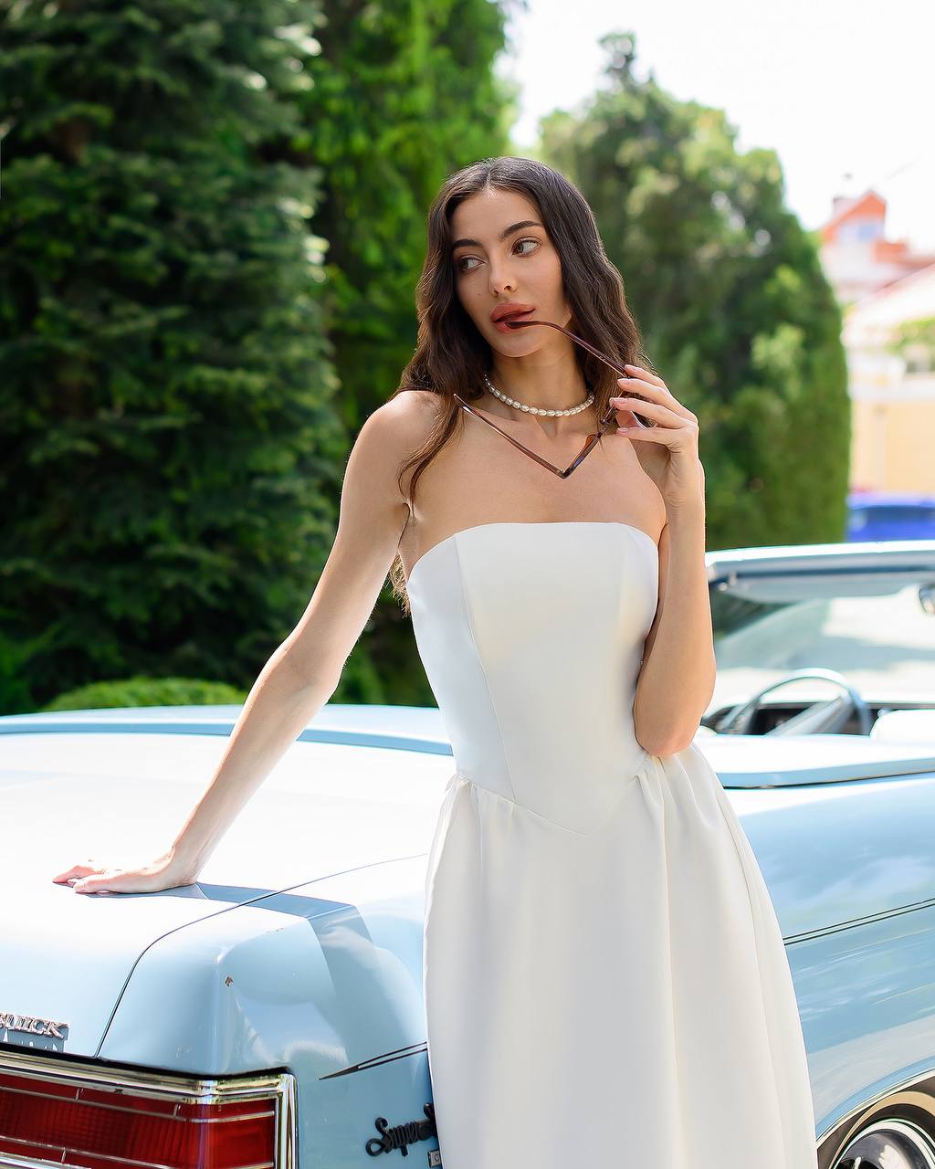 Woman in a white dress standing next to a vintage car with greenery in the background