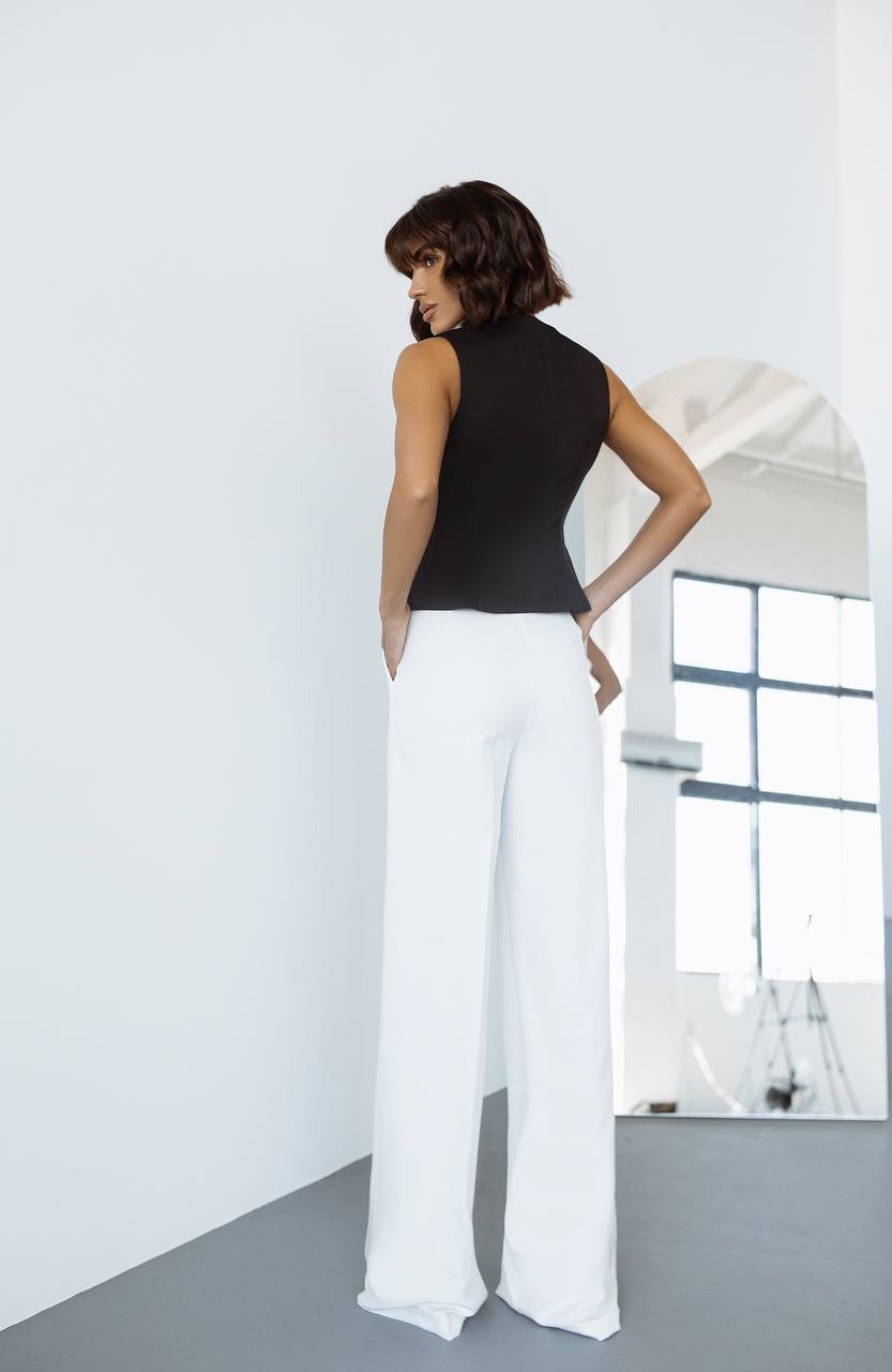 Woman wearing a black sleeveless top and white pants in a minimalistic room.