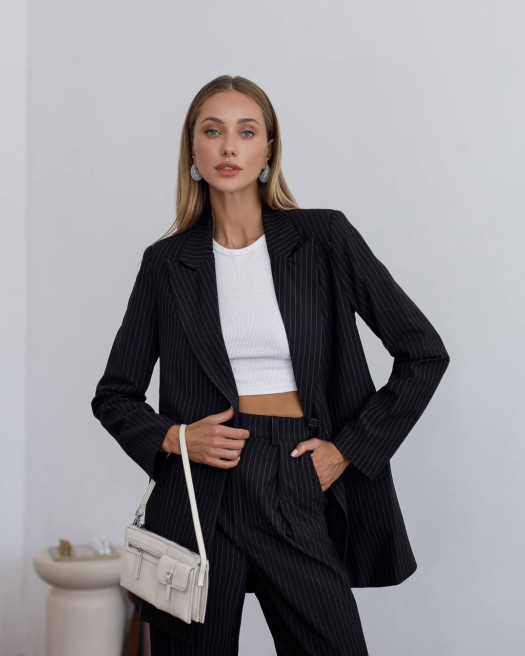 Woman wearing a black pinstripe suit with a white top, holding a beige handbag against a plain background.