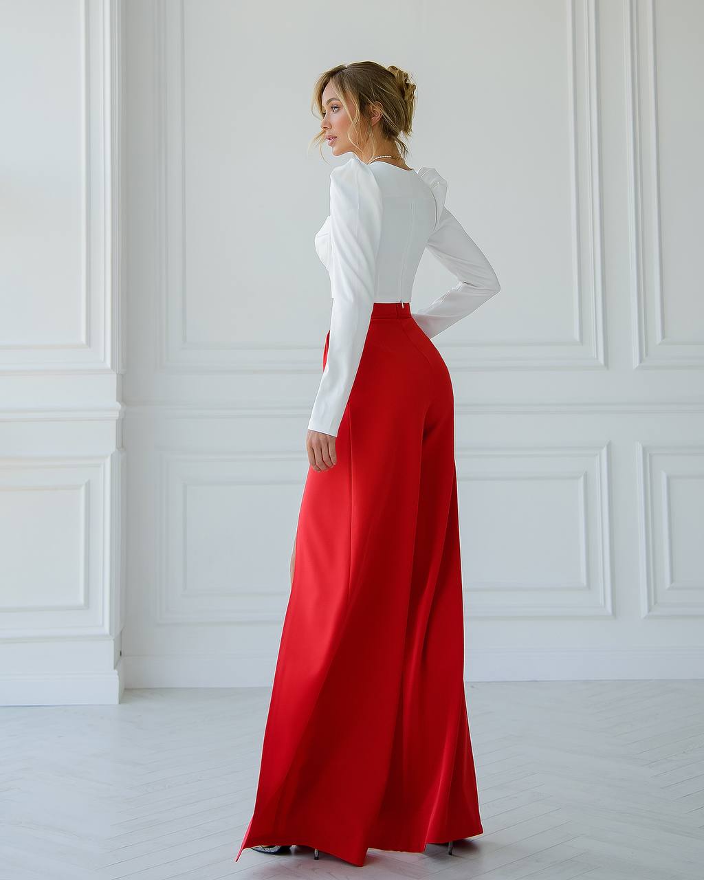 Woman wearing a white long-sleeve top and red high-waisted pants in a white room.