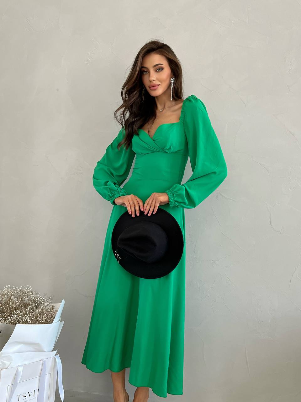 Woman in a green dress holding a black hat against a neutral background
