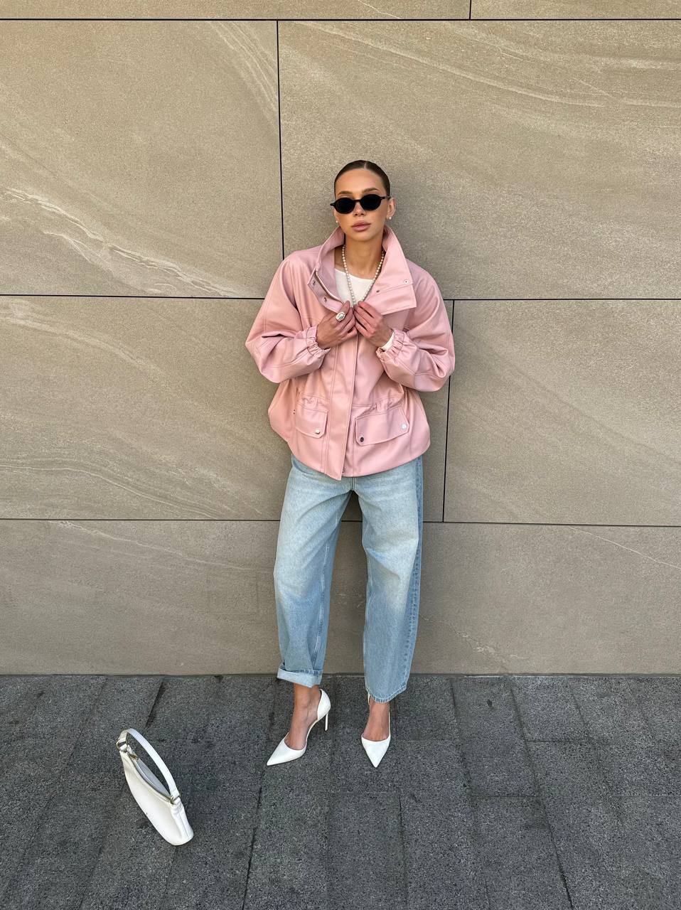 Person wearing a pink jacket and light blue jeans standing against a beige wall.