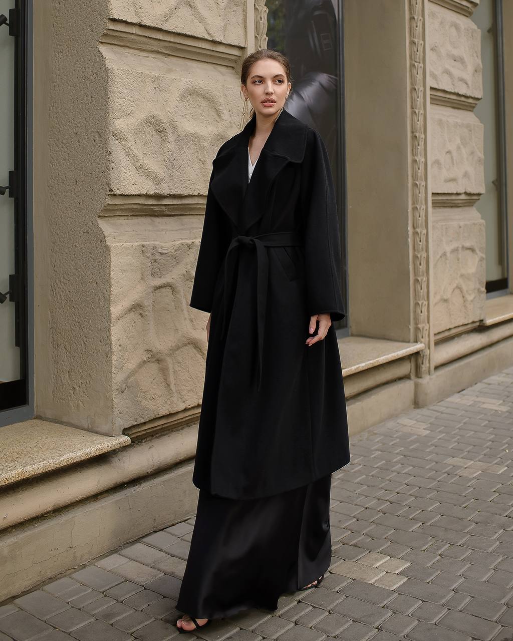 Woman wearing a long black coat standing on a street with stone building facades.