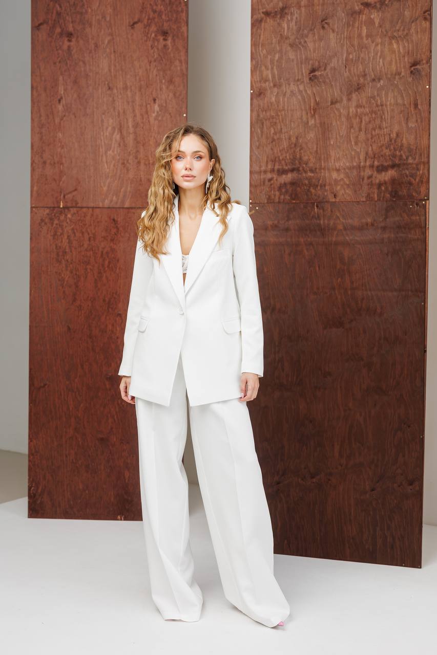 Woman in a white suit standing against a wooden wall.
