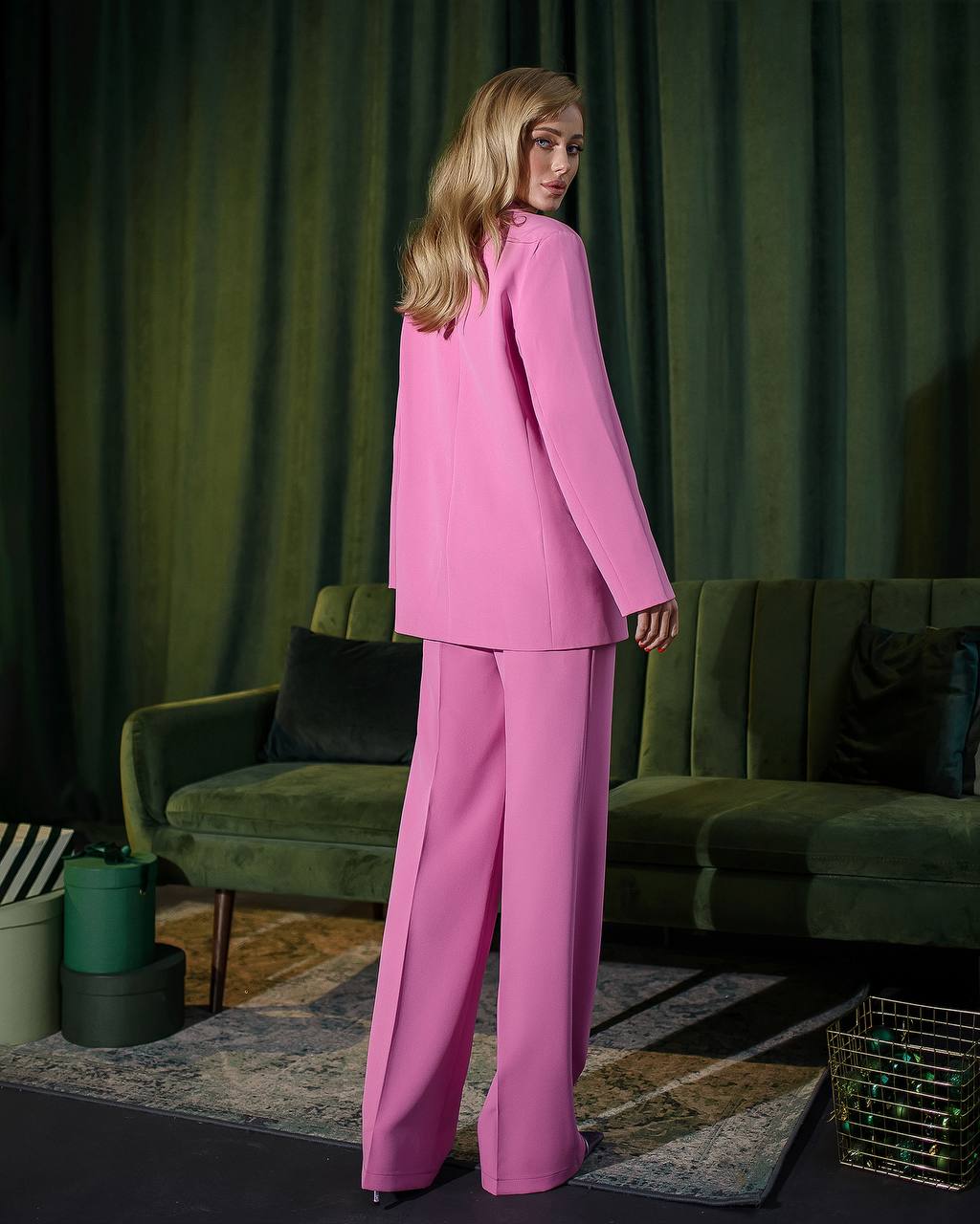 Woman in a pink suit standing in a room with green curtains and a green sofa.