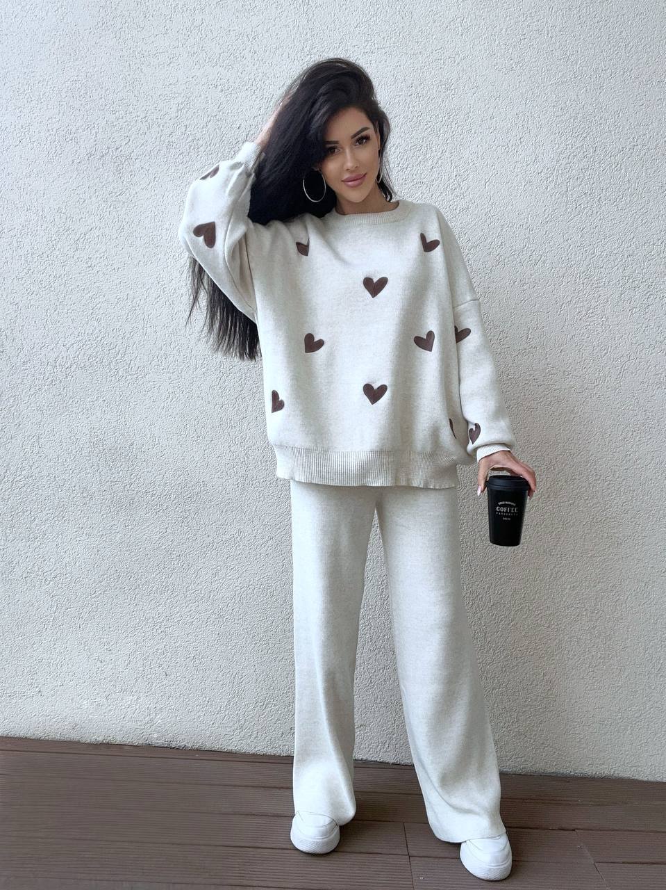Woman wearing a white sweater with heart patterns and matching pants, holding a black cup against a plain background.