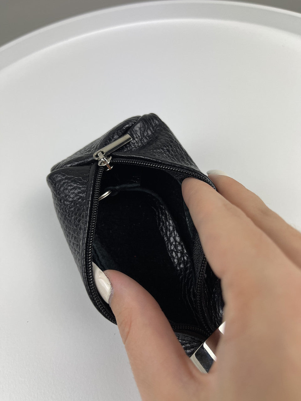 A close-up of a black textured leather zippered pouch being opened by a hand with white manicured nails, set against a clean white background.