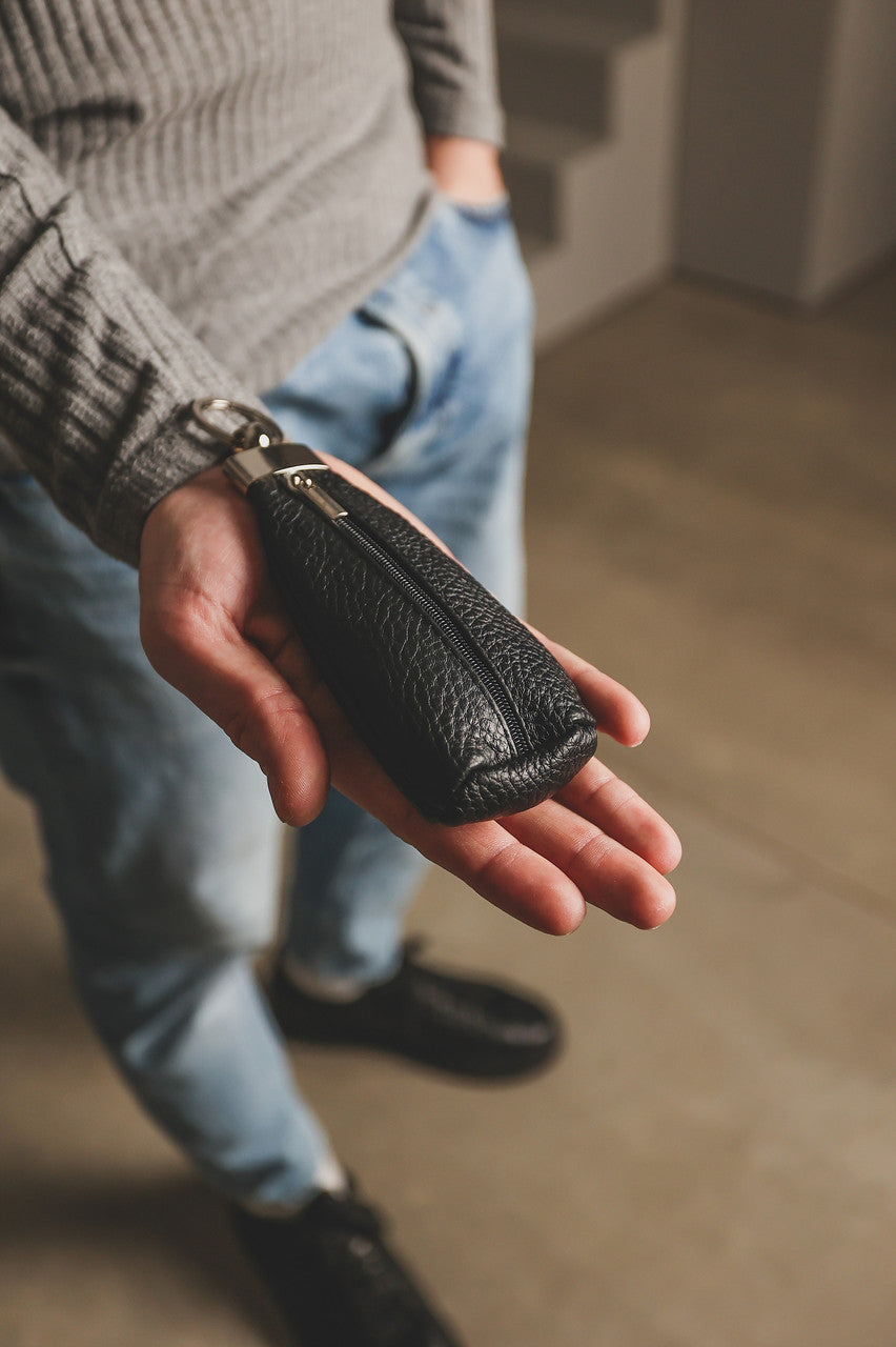 A close-up of a person's hand holding a small black pebbled leather zippered pouch or wallet, with a gray ribbed sweater, blue jeans, and black shoes visible in a softly lit indoor setting.