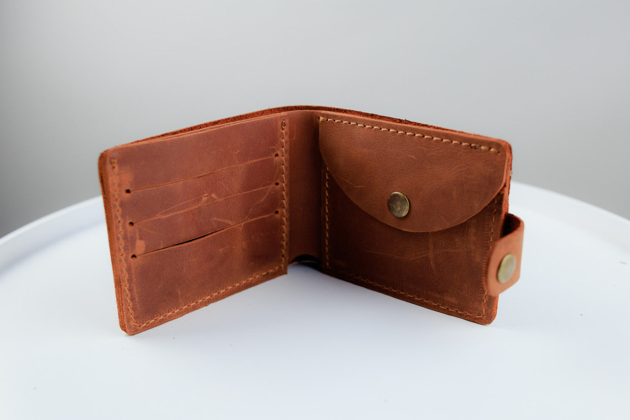 A brown leather bi-fold wallet shown open, displaying its interior card slots and a front pocket with a snap button closure, set against a neutral gray and white background.