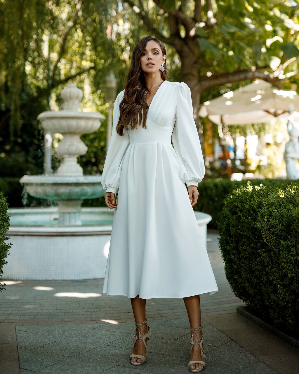 Woman in a white dress standing outdoors with a fountain and greenery in the background