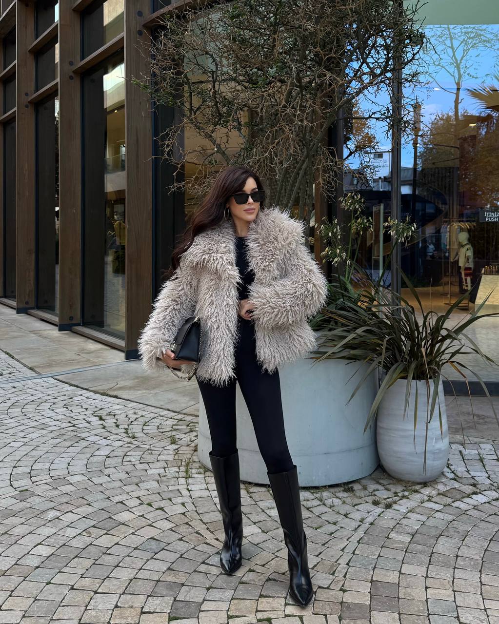 Woman in a fur coat standing on a cobblestone street with plants and a building in the background.