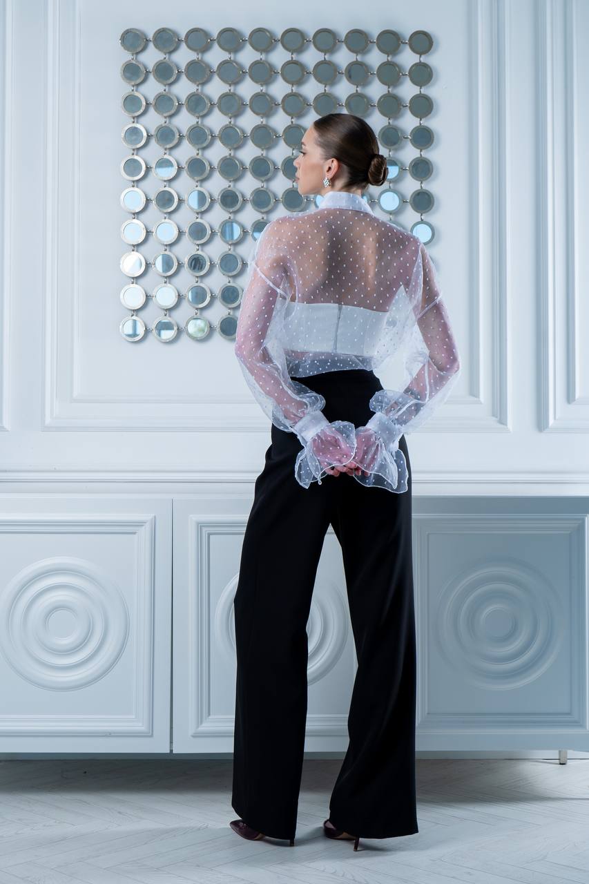 Person wearing a sheer top and black pants against a decorative wall.